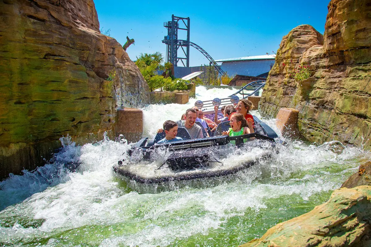 Des personnes sur un radeau aquatique s'ébattent dans un canal rocheux rempli d'eau sous un ciel bleu, avec des montagnes russes en arrière-plan.