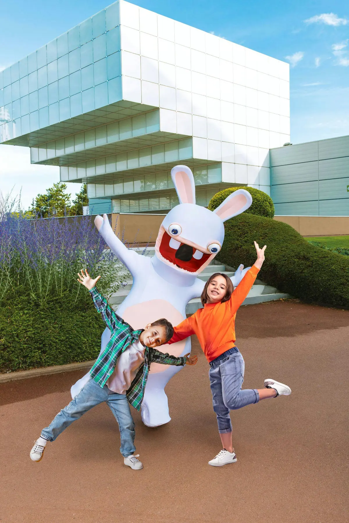 Deux enfants posent joyeusement avec une personne portant un grand costume de lapin de dessin animé, devant un bâtiment moderne et un jardin paysager.