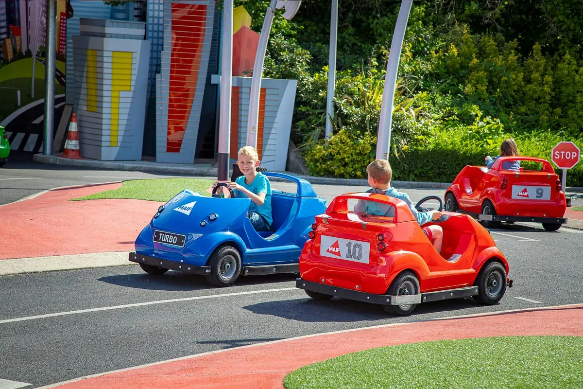 Deux enfants conduisent des mini-voitures, l'une bleue et l'autre rouge, sur une petite piste dans un parc d'attractions en plein air avec de la verdure.