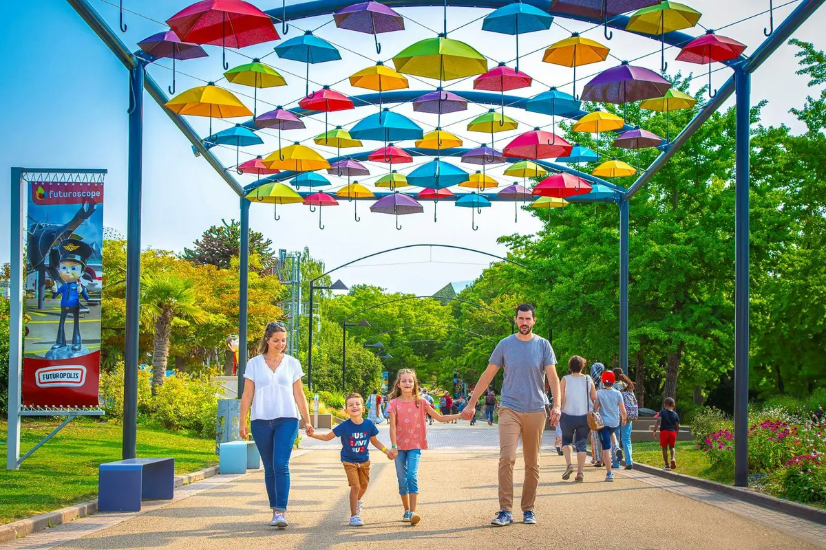 Une famille de quatre personnes se promène main dans la main sous un auvent de parapluies colorés dans un parc, avec de la verdure et d'autres visiteurs en arrière-plan.