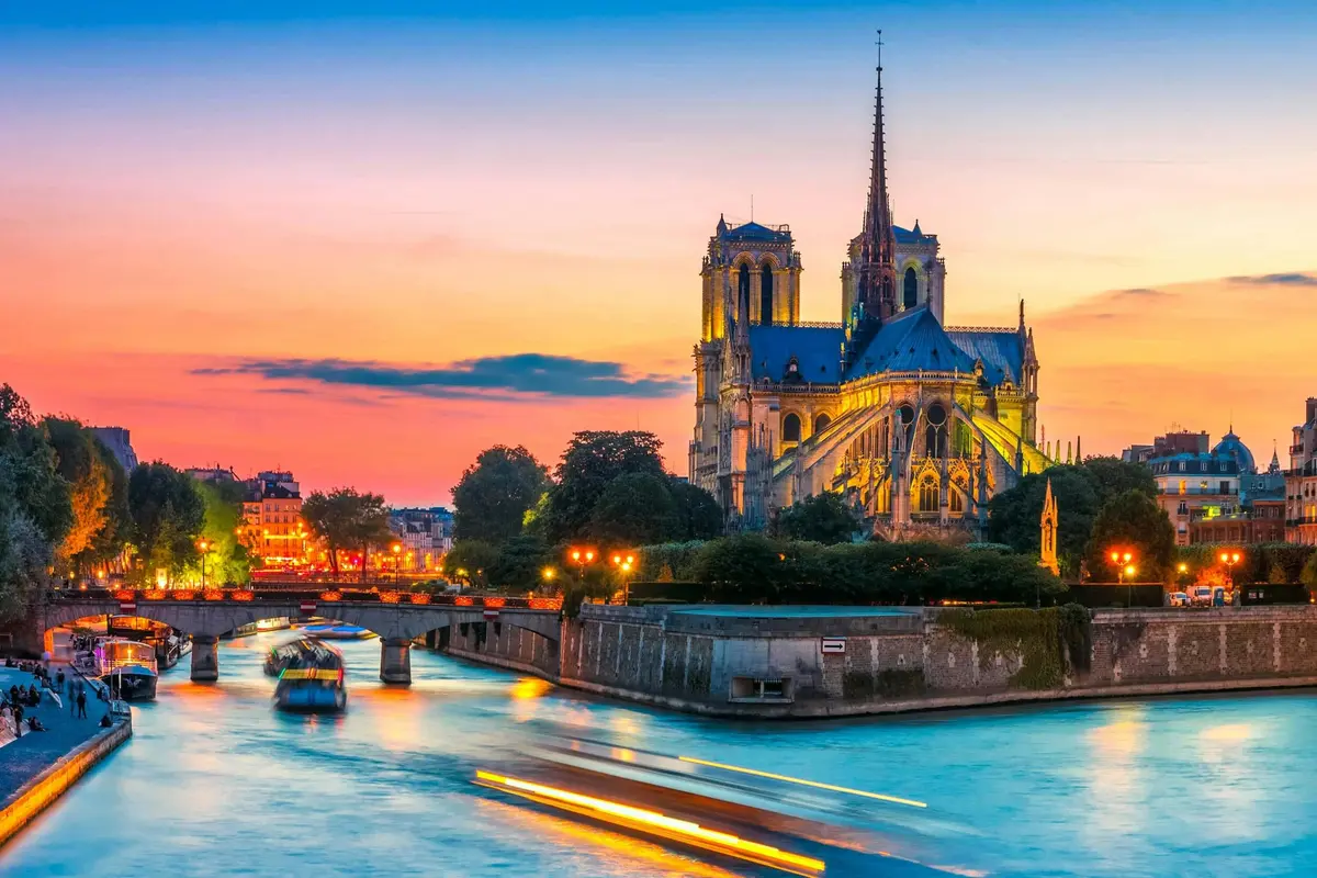 Cathédrale Notre-Dame au coucher du soleil avec une rivière au premier plan, des bateaux illuminés et un paysage urbain à l'arrière-plan.