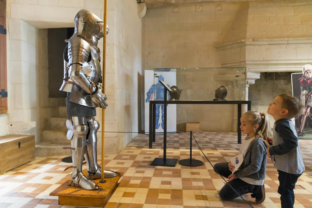 Deux enfants observent des armures médiévales exposées dans une salle de musée aux murs de pierre et au sol en damier.