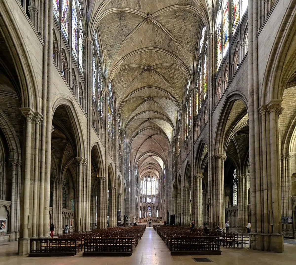 Un vaste intérieur de cathédrale avec des vitraux, des plafonds voûtés et des rangées de chaises en bois. Quelques personnes sont présentes.