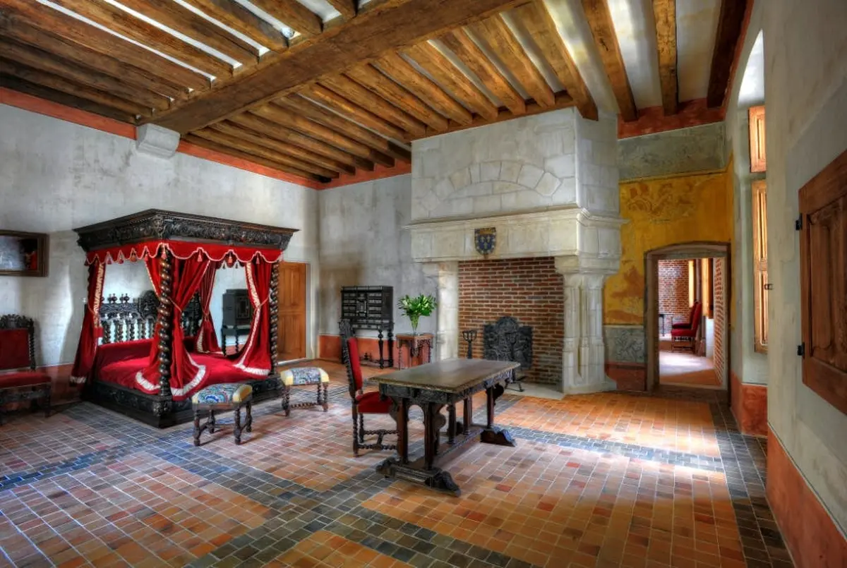 Une grande pièce avec un plafond en bois, une cheminée en pierre, un lit orné avec des rideaux rouges, une table en bois, des chaises et un sol carrelé.