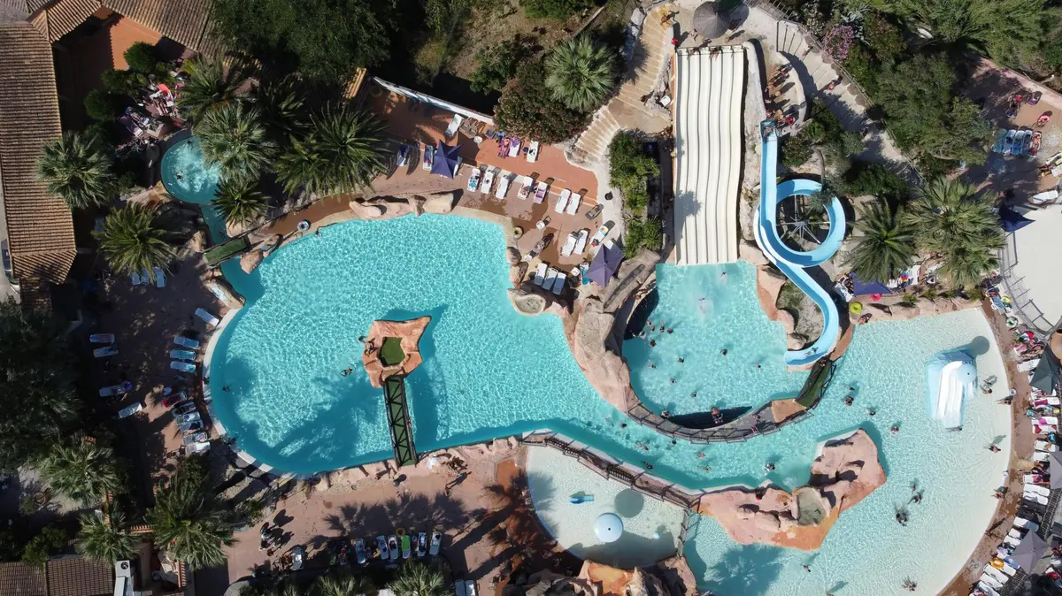 Vue aérienne d'un parc aquatique avec piscines, toboggans et chaises longues, entouré de végétation et de sentiers.