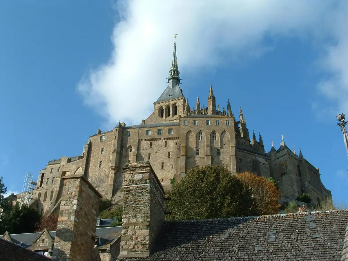 Grand château de pierre avec flèches et tourelles, entouré d'arbres, vu d'en bas avec les toits au premier plan. Ciel bleu avec des nuages au-dessus.