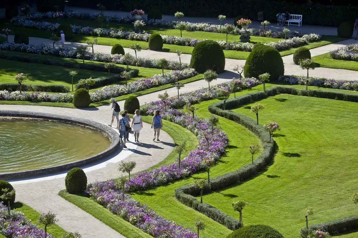 Vue aérienne de personnes marchant dans un jardin entretenu avec des parterres de fleurs, des haies et une fontaine circulaire.