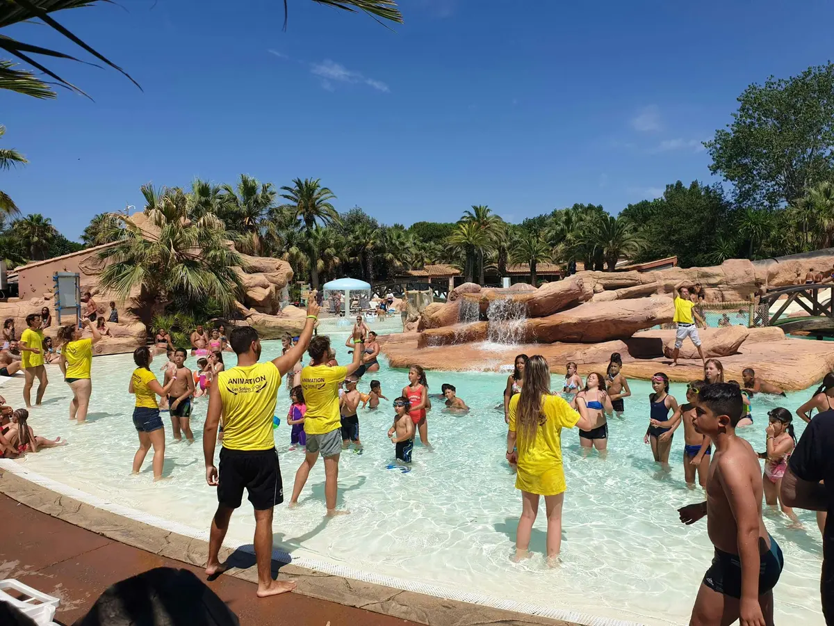 Des personnes s'amusent dans une piscine peu profonde entourée de palmiers, de rochers et de chutes d'eau sous un ciel bleu ensoleillé.