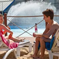 Une femme se prélasse sur un transat au bord d'une piscine, tandis qu'un homme est assis à proximité. Une personne en maillot de bain passe à côté d'elle et une fontaine projette de l'eau en arrière-plan. DR