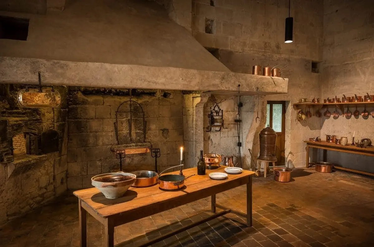 Une cuisine rustique, à l'ancienne, avec une grande cheminée en pierre, une table en bois, des casseroles en cuivre et divers ustensiles de cuisine.