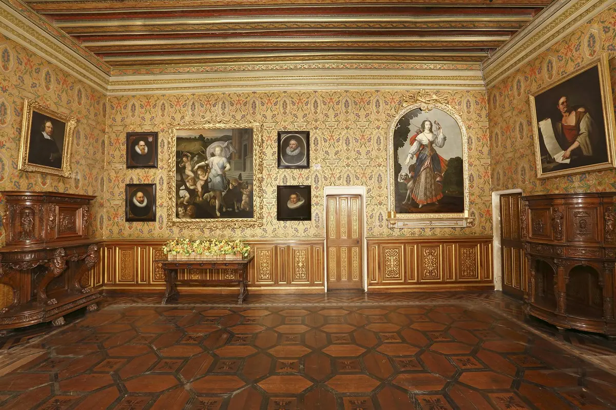 Une pièce richement décorée avec du papier peint orné, des portraits au mur et une table en bois ornée de fleurs.