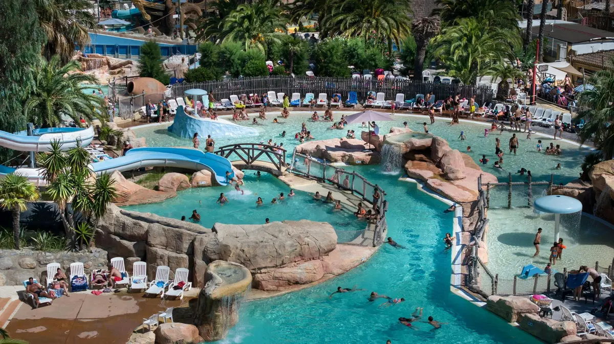 Un parc aquatique avec des piscines, des toboggans, des structures rocheuses et des gens qui se baignent et se détendent sur des chaises par temps ensoleillé.