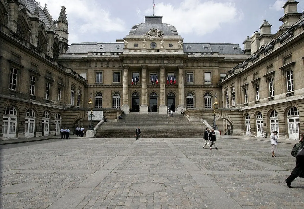 Un bâtiment historique avec des drapeaux, de grands escaliers et des gens qui marchent dans une cour spacieuse sous un ciel partiellement nuageux.