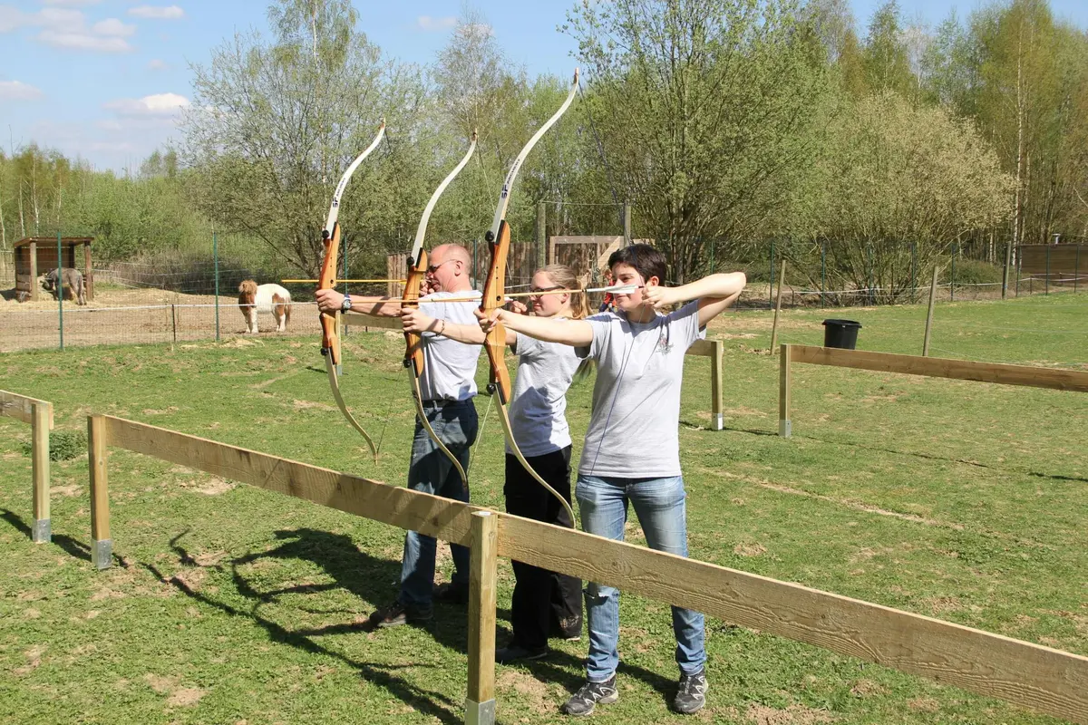 Trois personnes visant des arcs lors d'un entraînement au tir à l'arc en plein air, avec des arbres et une clôture en arrière-plan.