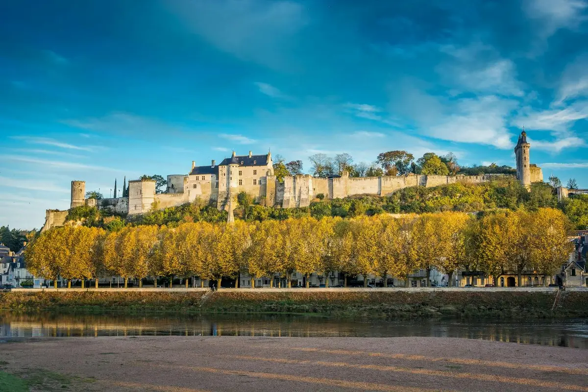 Un château historique avec des arbres jaunes d'automne au premier plan, sur fond de ciel bleu, et une rive en bas.