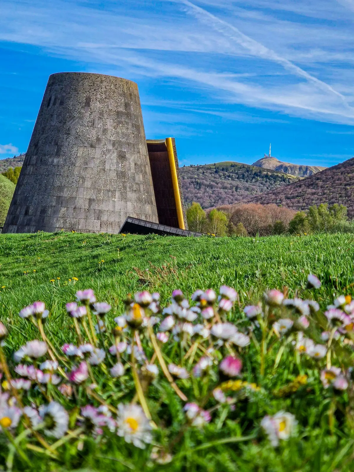 Une structure en pierre sur un champ herbeux avec des fleurs sauvages au premier plan et des collines avec une tour à l'arrière-plan sous un ciel bleu.