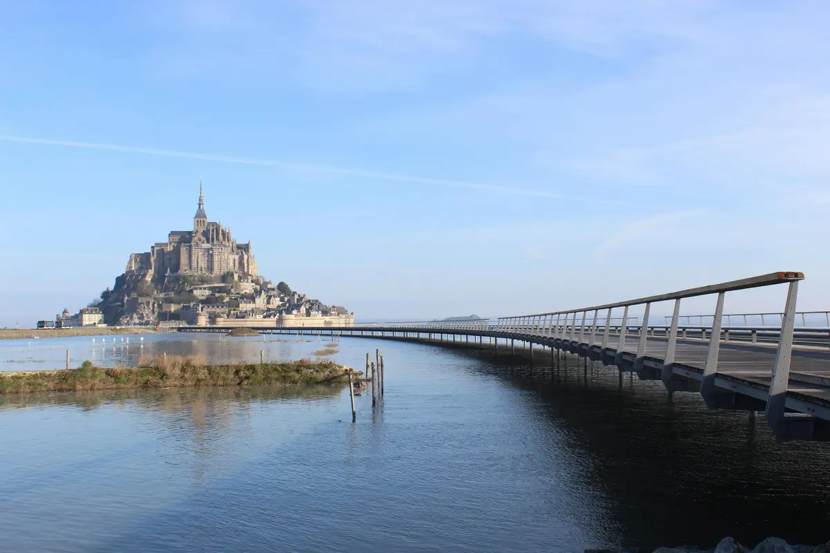 Un pont incurvé mène à une île historique dotée d'une structure haute et pointue, entourée d'une eau calme sous un ciel bleu clair.