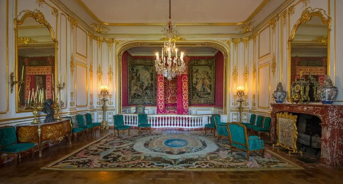 Pièce élaborée, ornée de détails dorés, de lustres, de rideaux rouges, de tapisseries, de chaises sarcelles et d'un grand tapis complexe.
