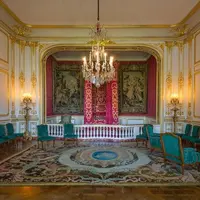 Pièce élaborée, ornée de détails dorés, de lustres, de rideaux rouges, de tapisseries, de chaises sarcelles et d'un grand tapis complexe. DR