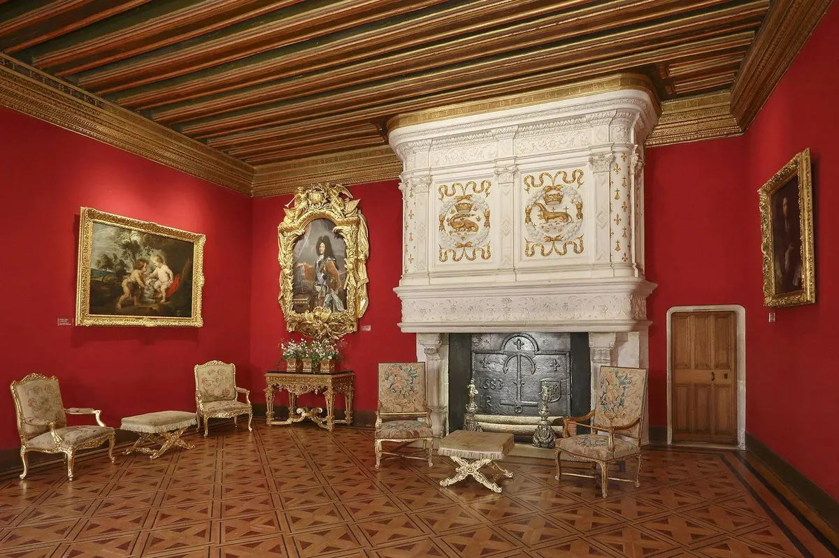Chambre élégante avec des murs rouges, un plafond orné, une grande cheminée décorative, des meubles anciens et des œuvres d'art encadrées.