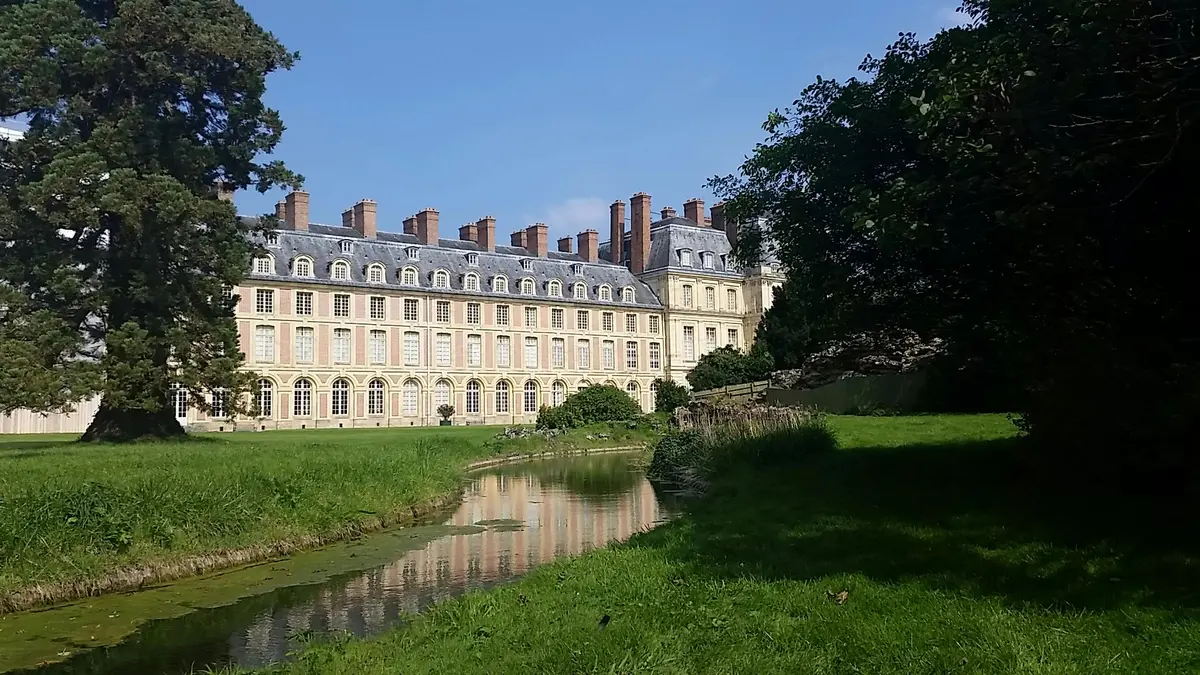 A historic building with numerous windows and chimneys, surrounded by greenery and a small reflective water stream.