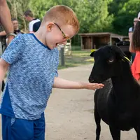 Un jeune garçon en chemise bleue et lunettes tend la main à une chèvre noire tandis qu'un adulte se tient à proximité ; d'autres personnes sont à l'arrière-plan. DR