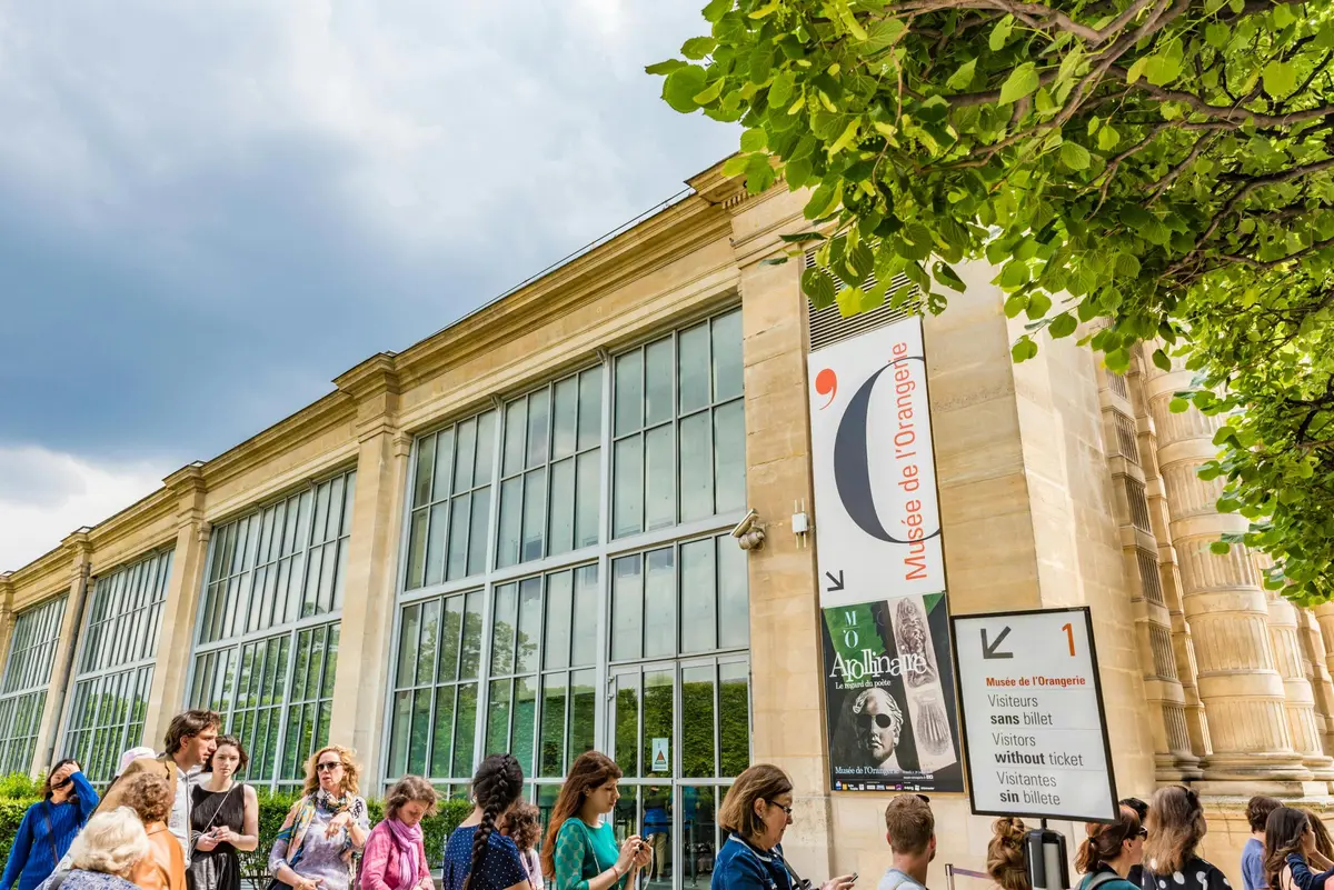Un groupe de personnes se tenant à l'extérieur d'un grand bâtiment avec de hautes fenêtres en verre et une affiche d'exposition sur le mur ; ciel couvert au-dessus.