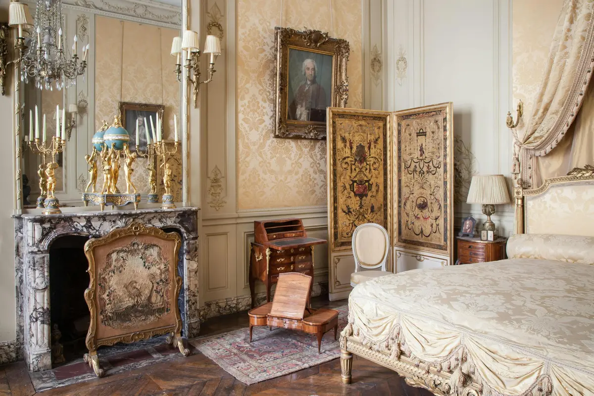 Une chambre à coucher ornée d'un lit luxueux, d'une cloison complexe, de meubles anciens, d'une cheminée en marbre et d'un portrait au mur.