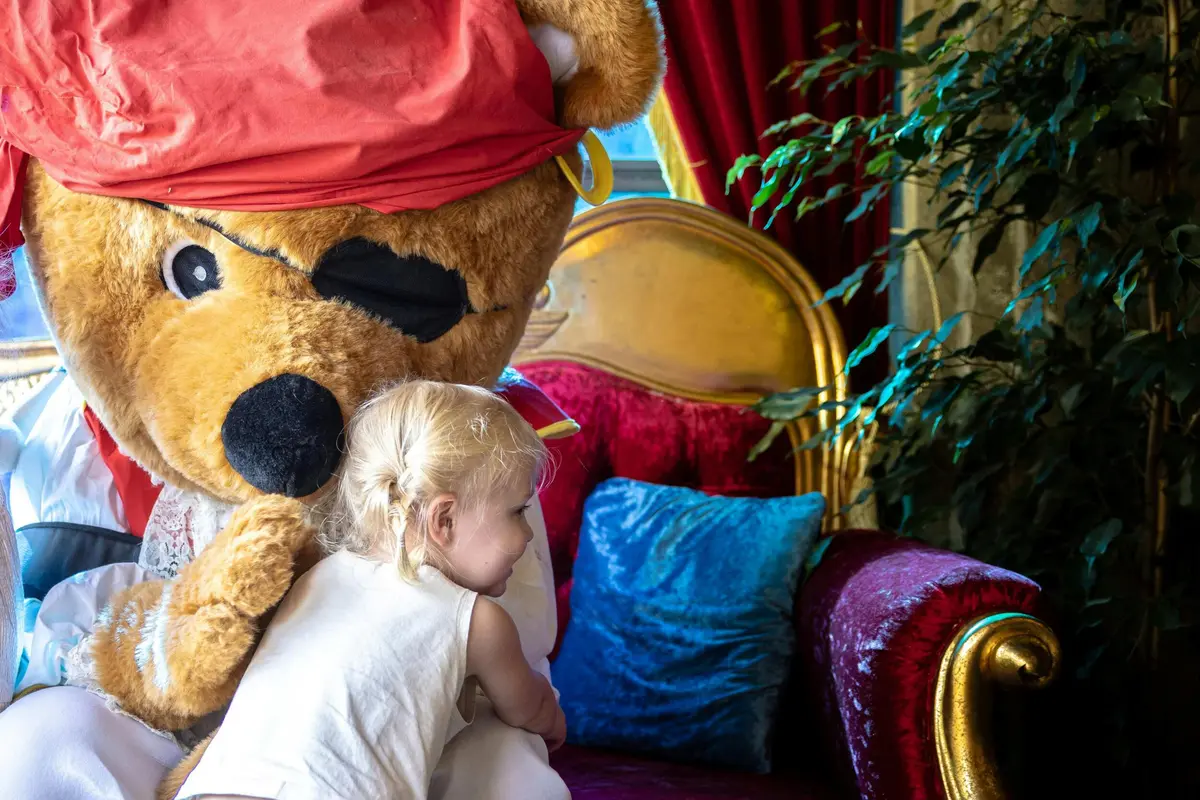 Un enfant enlace une personne déguisée en ours, assise sur une chaise en velours rouge, avec des coussins décoratifs et une plante feuillue à proximité.