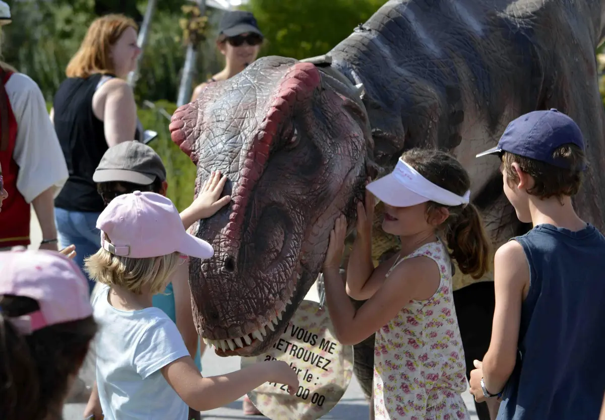 Des enfants portant des chapeaux interagissent avec une marionnette de dinosaure actionnée par une personne. Des adultes se tiennent debout à l'arrière-plan.