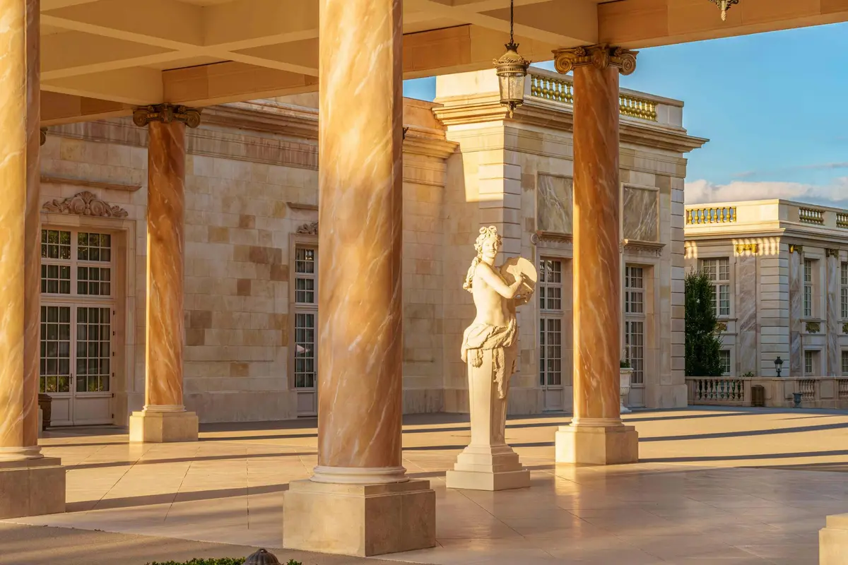 Une statue de marbre se dresse entre des colonnes dans une cour de style classique éclairée par le soleil et dotée d'une architecture en pierre ornée.