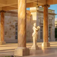 Une statue de marbre se dresse entre des colonnes dans une cour de style classique éclairée par le soleil et dotée d'une architecture en pierre ornée. DR
