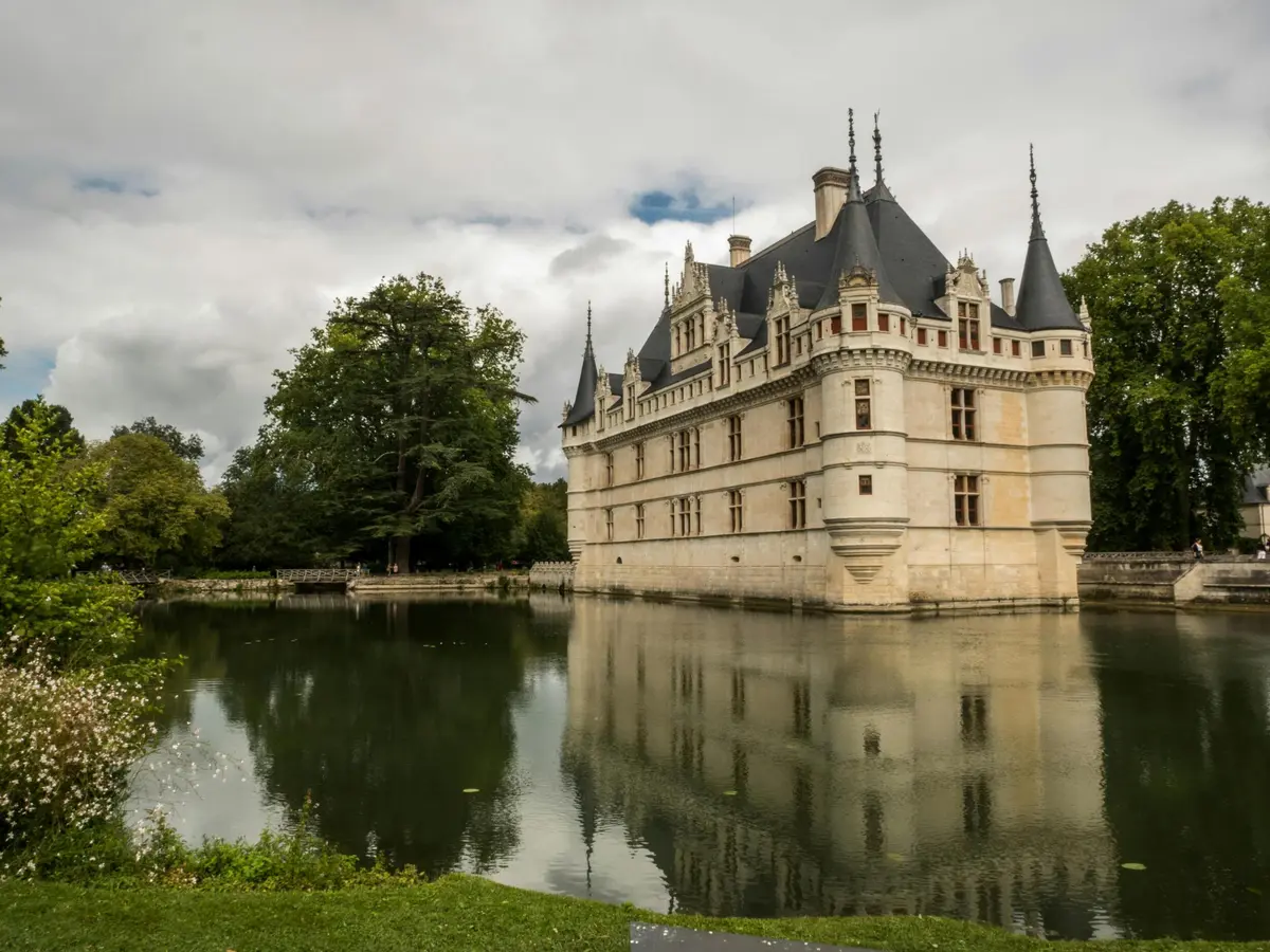 Un château historique en pierre avec des tours pointues se reflétant dans un lac calme, entouré d'arbres et de verdure sous un ciel nuageux.