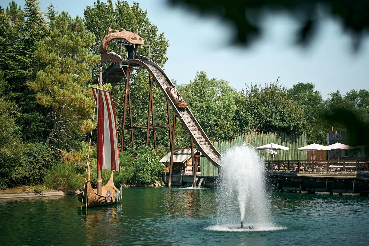 Manège aquatique avec une chute abrupte dans un bassin, un bateau viking avec une voile, et une fontaine au premier plan entourée de verdure.