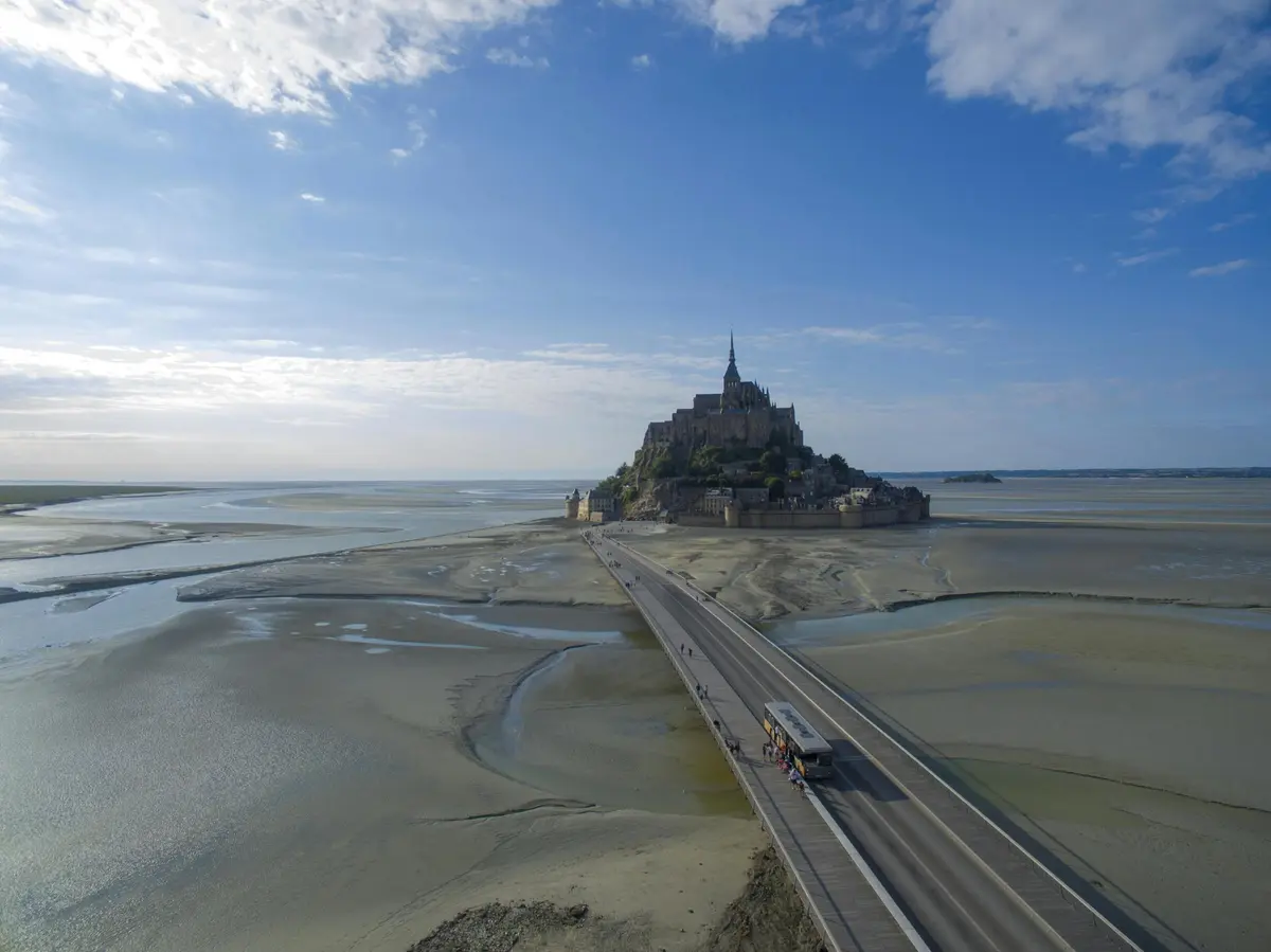 Un chemin de pierre mène à une abbaye insulaire historique entourée de bancs de sable à marée, sous un ciel bleu parsemé de nuages.