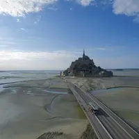 Un chemin de pierre mène à une abbaye insulaire historique entourée de bancs de sable à marée, sous un ciel bleu parsemé de nuages. &copy; Ryan R Zhao
