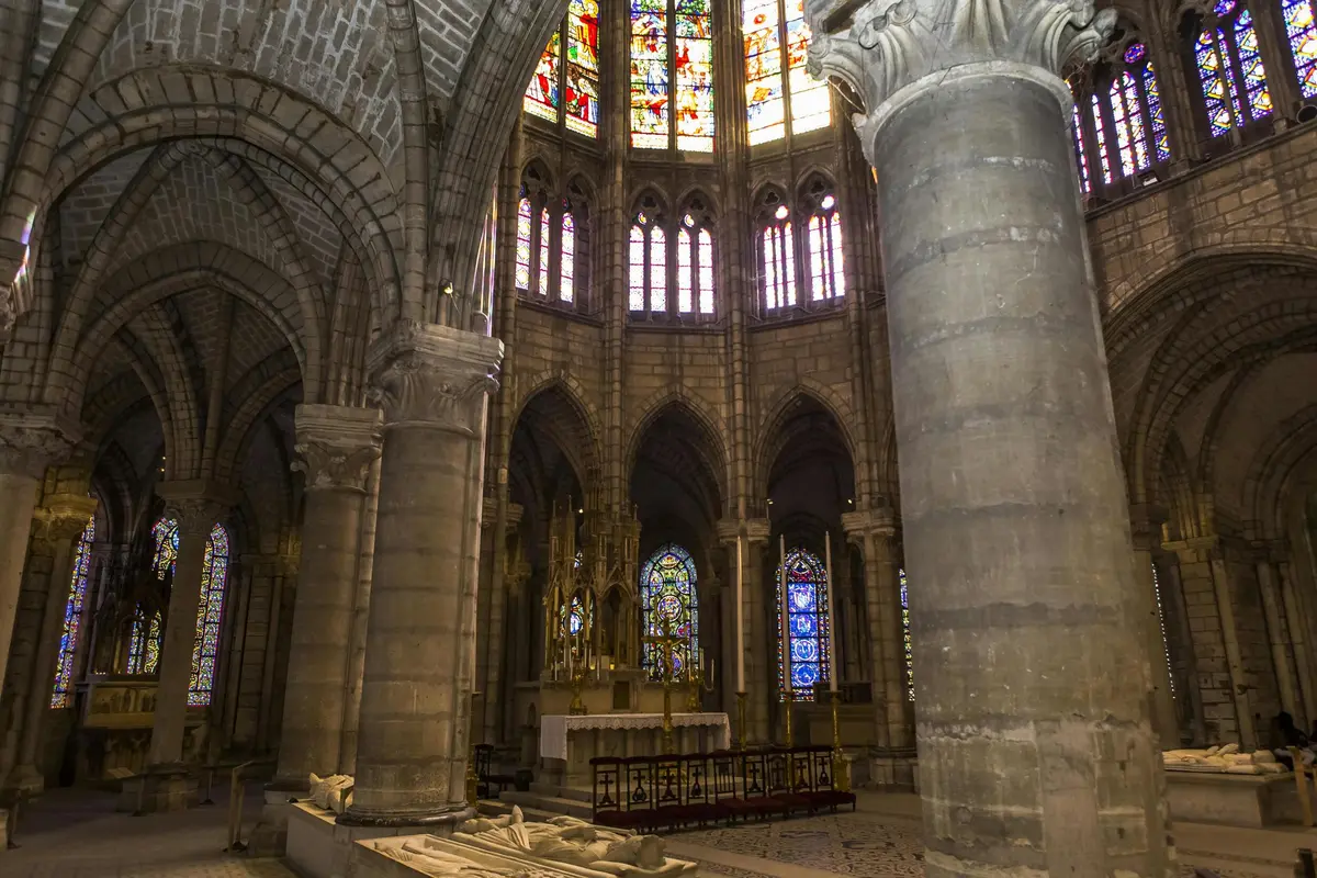 Intérieur de cathédrale gothique avec de grandes colonnes de pierre, des vitraux complexes et un autel orné à l'arrière-plan.