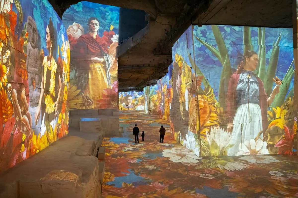 Une installation artistique immersive avec des projections vibrantes d'images inspirées de Frida Kahlo sur les murs et les sols, les visiteurs se promenant dans l'espace.