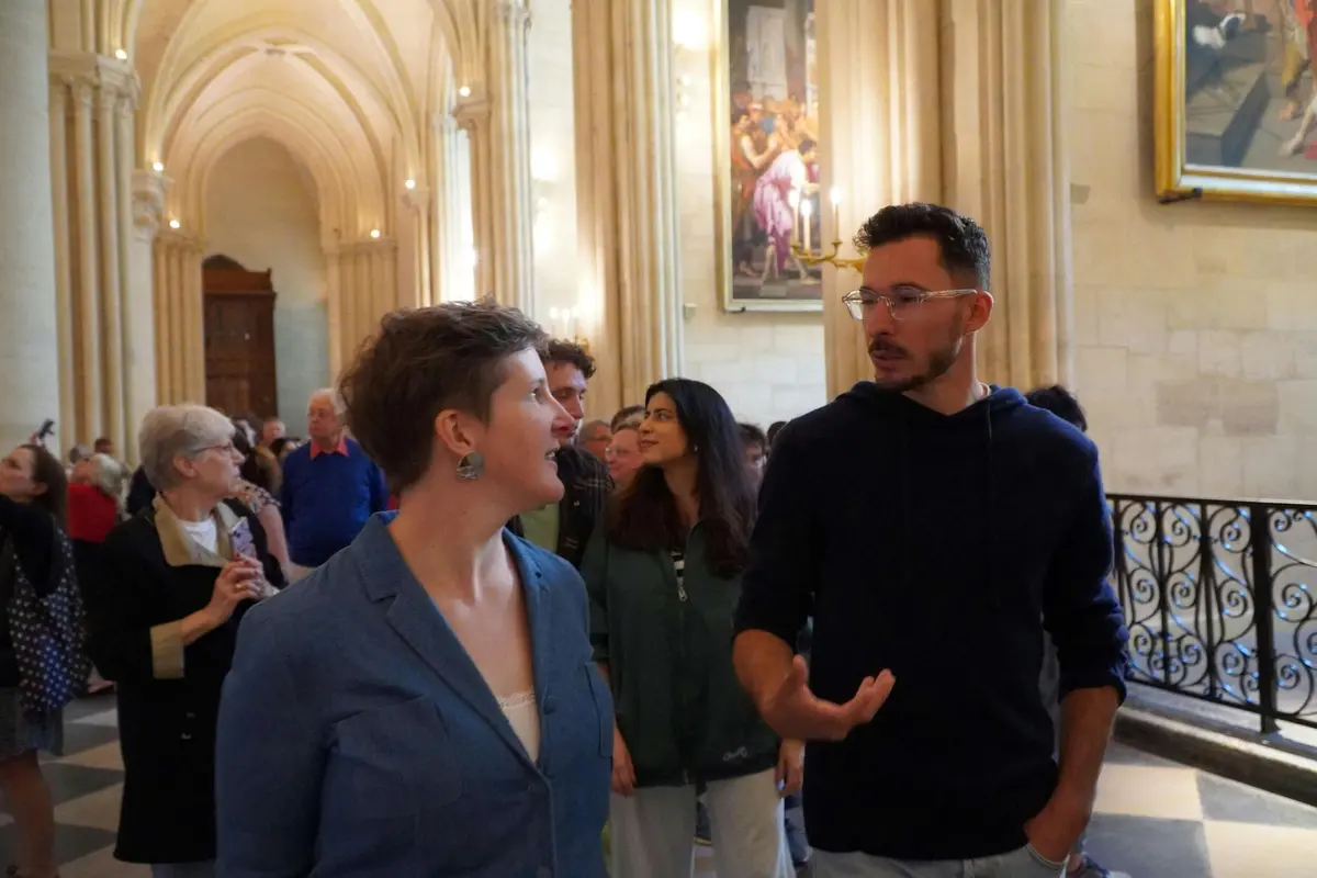 Des personnes discutent à l'intérieur d'un bâtiment historique avec des colonnes ornées, des œuvres d'art sur les murs et une balustrade décorative à l'arrière-plan.