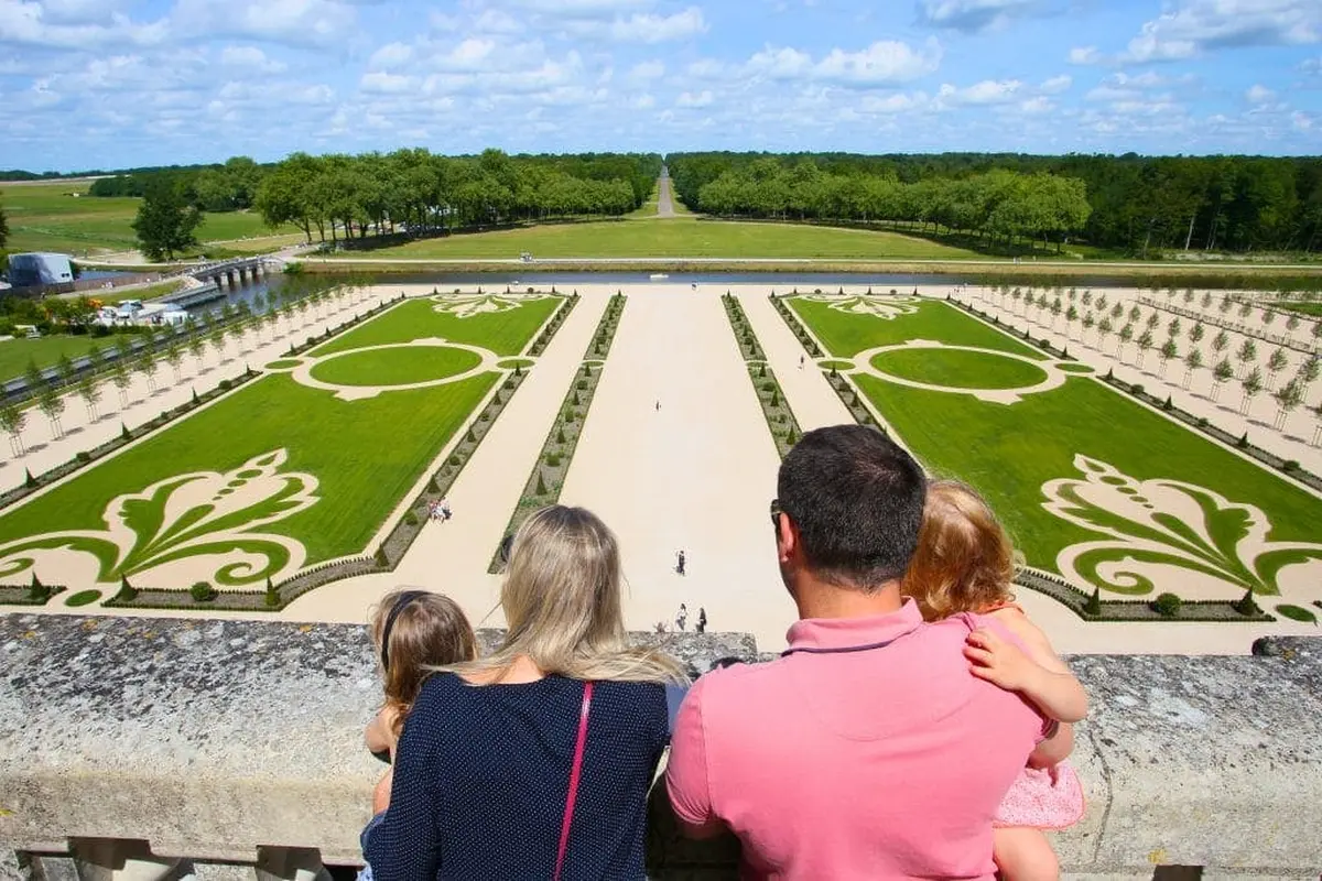 Une famille de quatre personnes contemple des jardins méticuleusement conçus, avec des allées symétriques et des pelouses vertes ornées de motifs décoratifs.