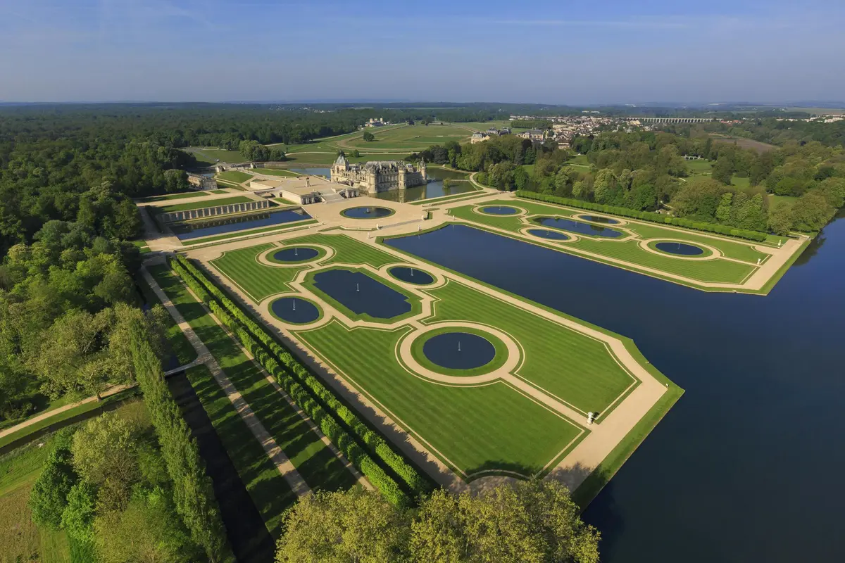 Vue aérienne d'un palais avec des jardins symétriques, de grands étangs et des pelouses entretenues, entouré d'arbres et de bâtiments éloignés.