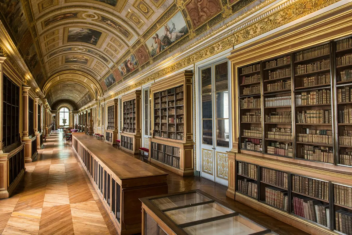 A grand library with a vaulted painted ceiling, wooden floors, bookshelves, and glass display cases along the corridor.
