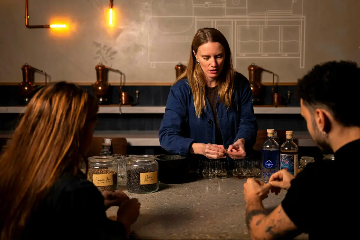 Une femme en chemise bleue prépare des articles sur une table avec des bocaux et des bouteilles, tandis que deux personnes observent dans un cadre confortable.