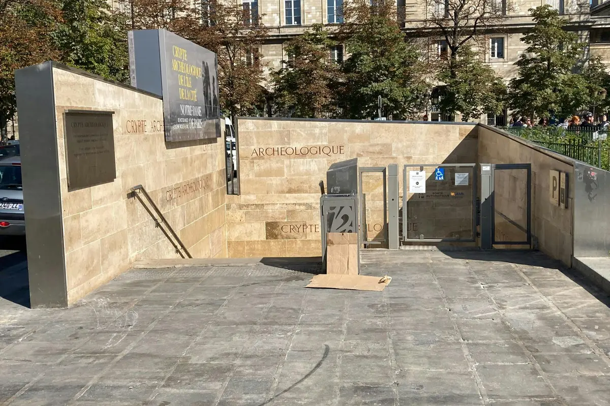 Entrée en pierre d'une crypte archéologique, avec signalisation et escaliers. Des arbres et un bâtiment sont visibles à l'arrière-plan.
