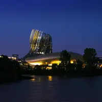 Bâtiment moderne illuminé au design incurvé près d'une rivière au crépuscule, entouré d'arbres et de lumières de la ville. DR