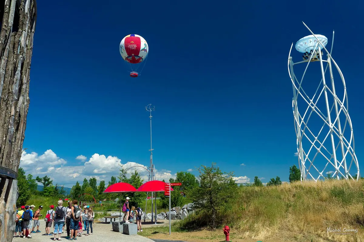 Des personnes se sont rassemblées dans un parc avec des parapluies rouges, une montgolfière attachée dans le ciel et une sculpture sur la droite dans un ciel bleu clair.