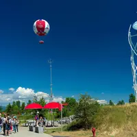 Des personnes se sont rassemblées dans un parc avec des parapluies rouges, une montgolfière attachée dans le ciel et une sculpture sur la droite dans un ciel bleu clair. DR
