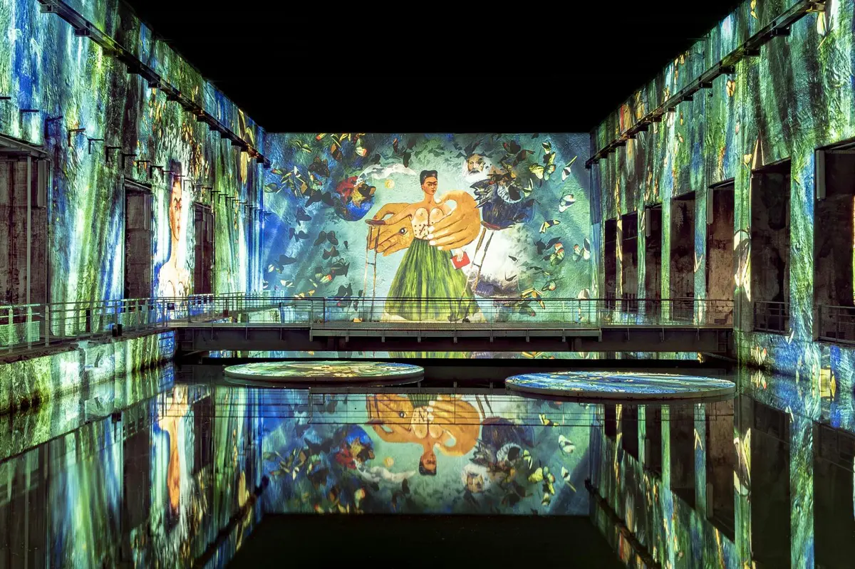 Une grande exposition intérieure avec des projections murales vibrantes présentant des œuvres d'art, dont une figure en vert, se reflétant sur l'eau en contrebas.