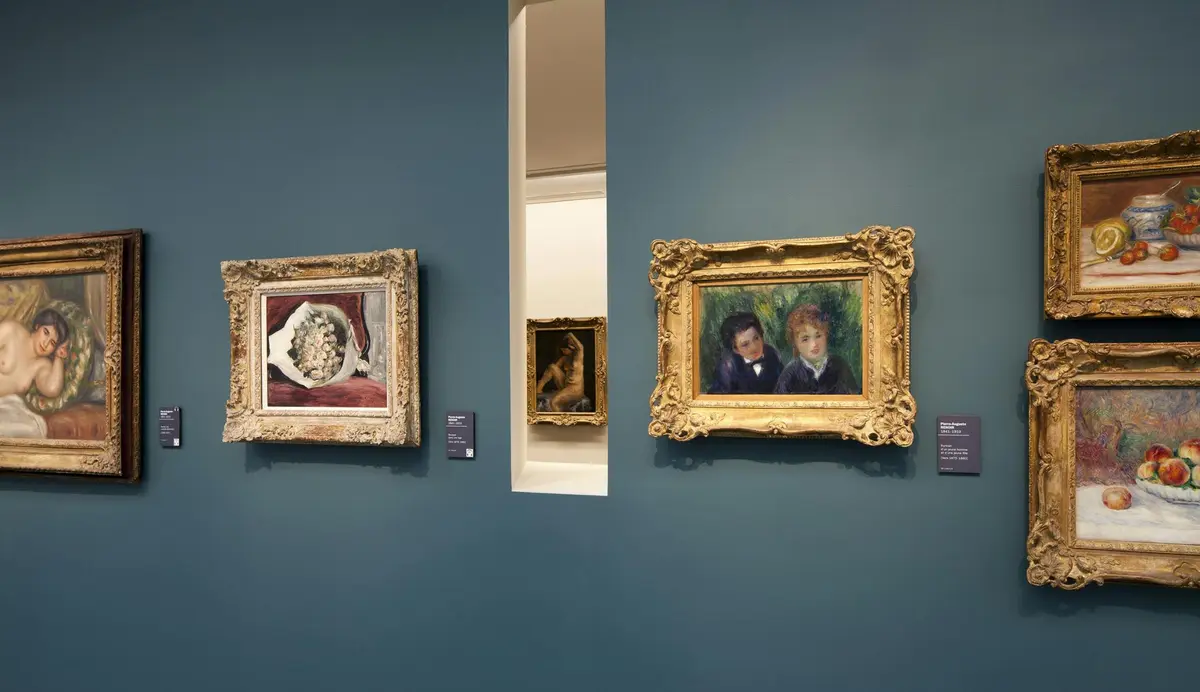 Des peintures dans des cadres ornés sont accrochées au mur d'une galerie sarcelle, présentant des portraits et des natures mortes, avec une petite sculpture visible à l'arrière-plan.