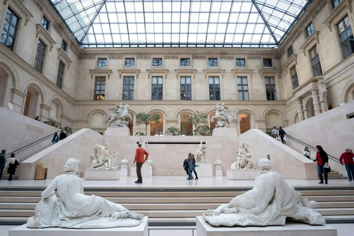 Une salle de musée avec des sculptures, des escaliers et des gens qui marchent, sous un plafond de verre et entourée d'une architecture classique.